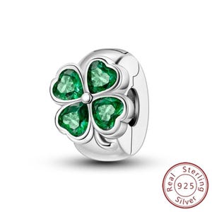 May include: A silver charm with a four-leaf clover design. Each heart-shaped leaf is filled with a vibrant green gemstone. The charm is round with a clasp and the words "Real Sterling 925 Silver" are printed on the bottom.