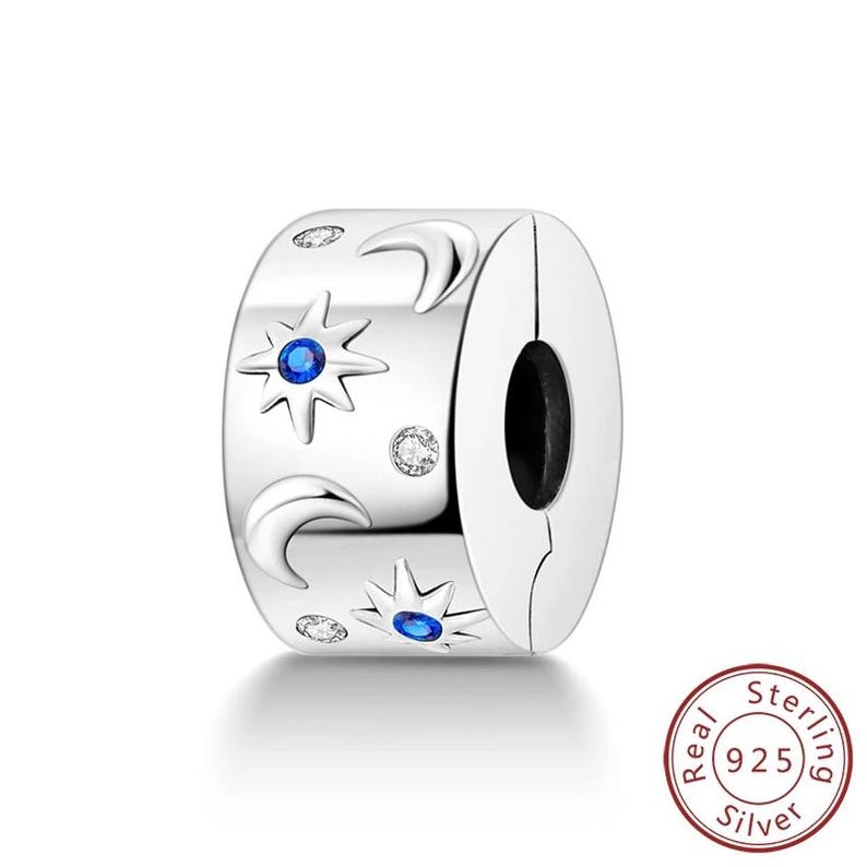 May include: A silver charm with a celestial design. The charm features crescent moons, starbursts with blue center stones, and small clear stones. The charm is marked with "Real 925 Silver".