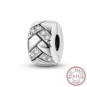 May include: A silver charm with a woven design, accented with clear, sparkling stones. The charm is a rectangular shape with a clasp on one side. The charm is made of 925 sterling silver.