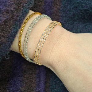 May include: Three beaded bracelets in gold, silver, and gold are stacked on a wrist. The bracelets are thin and delicate, with a textured appearance. The background is a dark, textured fabric, providing contrast to the jewelry.
