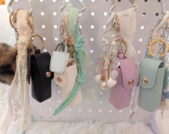 May include: A collection of keychains and accessories, including small leather pouches in black, white, pink, and mint green, adorned with gold hardware. Some keychains feature decorative elements like a fluffy pom-pom, beaded strands, and ribbons.