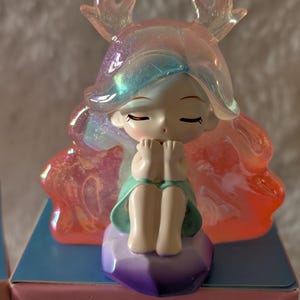 May include: A figurine of a seated girl with closed eyes, wearing a teal dress and a translucent headpiece with antler-like shapes. The figurine sits on a purple, geometric base. The background is a box with Chinese text and the words "FULL DISPLAY".