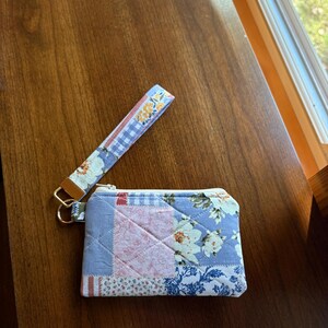 May include: A rectangular quilted fabric pouch featuring a patchwork design of blue, pink, and white floral and gingham patterns. The pouch includes a white zipper closure and a matching fabric wrist strap attached with a gold metal ring.