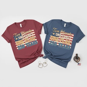 May include: Two short-sleeved t-shirts, one burgundy and one navy blue, each with the text "Two Hearts, One Flag, 250 Years" in a patriotic design. The shirts are knotted at the bottom. Accessories include heart-shaped earrings, a leather bracelet, and a small American flag.