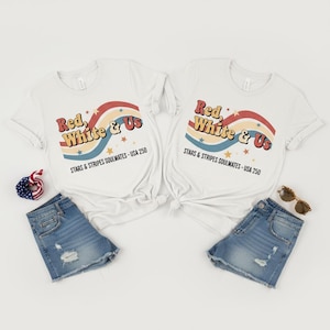 May include: Two white t-shirts with the text "Red, White & Us" in a retro font, above a wavy design in red, blue, and yellow. The shirts are paired with denim shorts, sunglasses, and a patriotic accessory, perfect for a Fourth of July celebration.