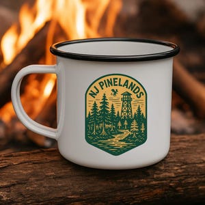 May include: White enamel camping mug with a black rim and handle. The mug features a green and yellow graphic with the text "NJ Pinelands" and a forest scene with a fire tower. The mug is sitting on a log.