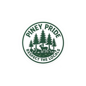 May include: A round sticker with a green and white design. The sticker features the text "PINEY PRIDE" and "RESPECT THE LOCALS" around a central image of pine trees, deer, rabbits, and a fox. The design is set against a white background.