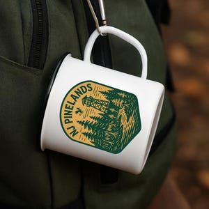 May include: White ceramic mug with a green and gold graphic of a forest scene and the text "NJ Pinelands." The mug is attached to a green backpack with a silver carabiner clip. Ideal for camping and outdoor enthusiasts.