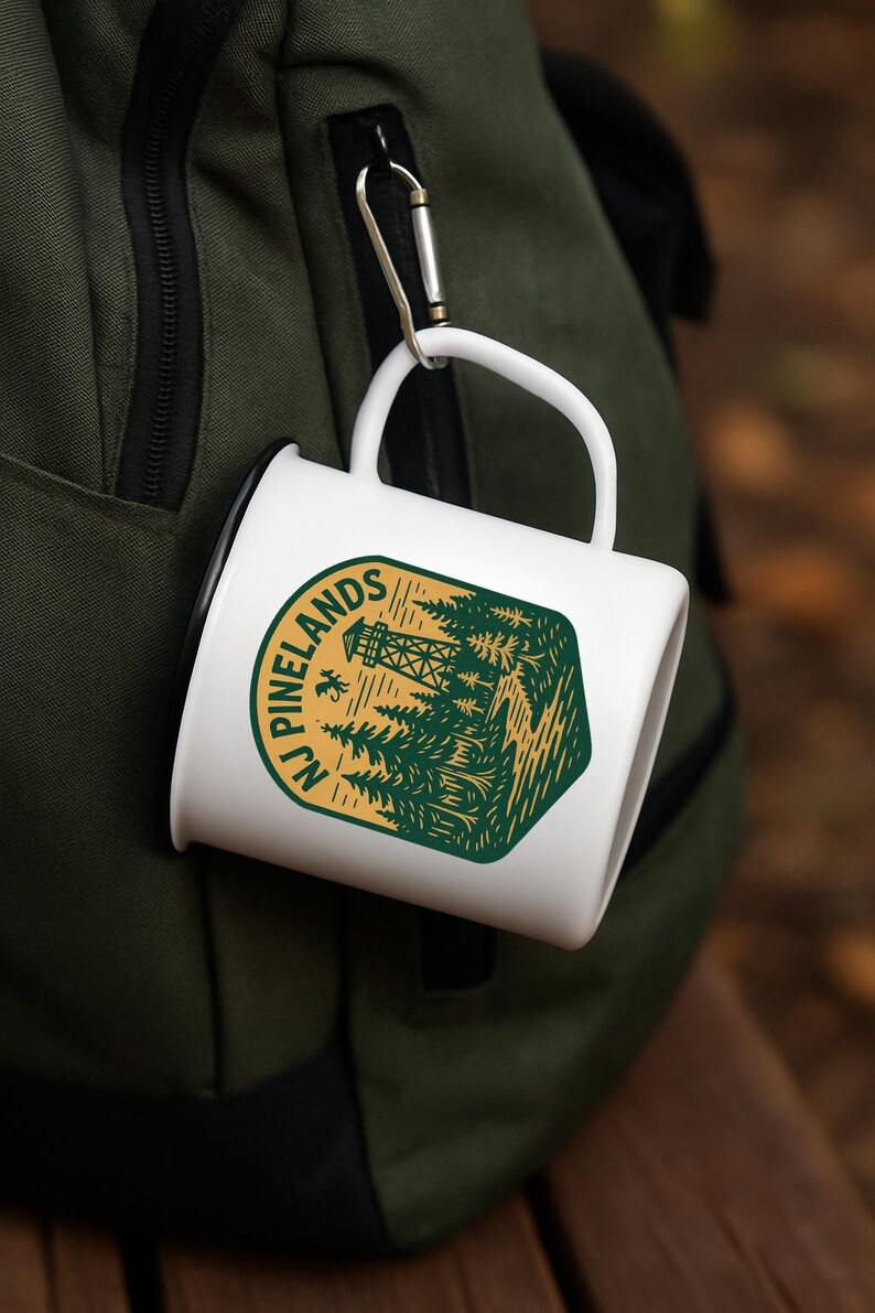 May include: White ceramic mug with a green and gold graphic of a forest scene and the text "NJ Pinelands." The mug is attached to a green backpack with a silver carabiner clip. Ideal for camping and outdoor enthusiasts.