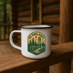 May include: White enamel mug with a black rim and handle. The mug features a green and yellow graphic with the text "NJ Pinelands", a forest scene, and a watchtower. The mug is sitting on a wooden surface.
