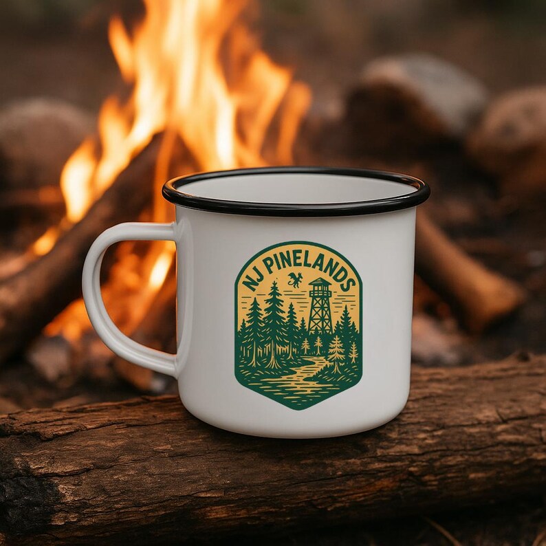 May include: White enamel camping mug with a black rim and handle. The mug features a green and yellow graphic with the text "NJ Pinelands" and a forest scene with a fire tower. The mug is sitting on a log.