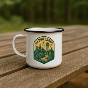 May include: White enamel mug with a black rim and handle. The mug features a green and yellow graphic of a forest scene with a watchtower and the text "NJ Pinelands". The mug is sitting on a wooden surface.