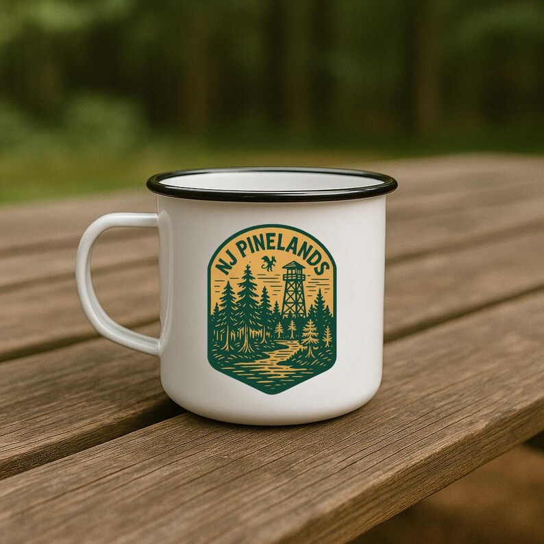 May include: White enamel mug with a black rim and handle. The mug features a green and yellow graphic of a forest scene with a watchtower and the text "NJ Pinelands". The mug is sitting on a wooden surface.