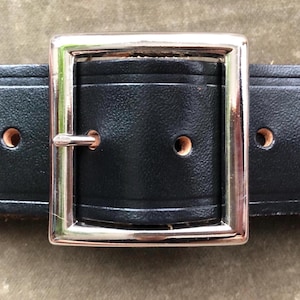 Può includere: Primo piano di una cintura in pelle nera con una fibbia rettangolare color argento. La cintura ha diversi fori distanziati uniformemente. La fibbia ha una cornice argentata e un perno argentato.