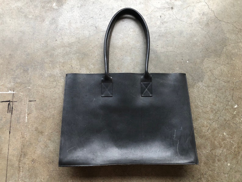 Puede incluir: Un bolso tote de cuero negro con dos asas superiores. El bolso tiene un dise&ntilde;o sencillo y est&aacute; hecho de un material de alta calidad.