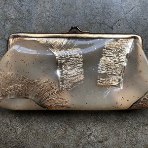 May include: A vintage gold-toned clutch purse with a clear plastic body and a gold glitter design. The clutch has a clasp closure.