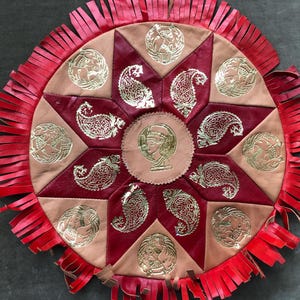 May include: A round, decorative leather piece with a red fringe. The center features a light brown circle with a gold profile. The surrounding sections are red and light brown, with gold paisley and medallion designs.