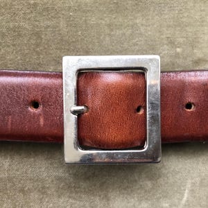 May include: Close-up of a brown leather belt with a silver square buckle. The belt has three holes and a rich, textured appearance. The buckle is centered, showcasing the belt's design.
