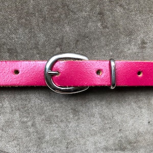 Könnte beinhalten: Ein rosa Ledergürtel mit einer silbernen Schnalle. Der Gürtel hat mehrere Löcher zum Einstellen der Größe.