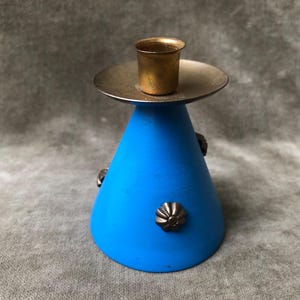 May include: A blue candlestick holder with a conical base and a brass candle cup. The holder has decorative metal accents and a silver-colored plate. The candlestick is a decorative home accent.
