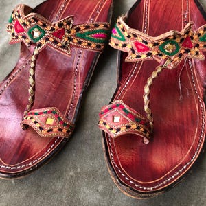 May include: A pair of handcrafted leather sandals with intricate embroidery. The sandals feature a rich brown color with decorative straps in red, green, and gold. The toe strap is a twisted gold cord.
