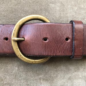 May include: Close-up of a brown leather belt with a large, round, brass buckle. The belt has several holes for adjustment and a loop to secure the end. The leather shows signs of wear.
