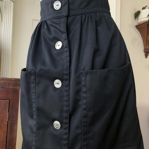 May include: A black skirt with large front pockets and a button-up closure. The skirt has a high waist and is made of a solid, dark fabric. The buttons are white and round. The skirt is displayed on a mannequin.