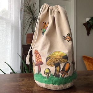 May include: A beige drawstring bag with a hand-painted design of colorful mushrooms and butterflies. The mushrooms are in shades of yellow, brown, and orange, set against a green grassy base. Two butterflies are also painted on the bag.