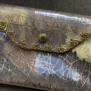 May include: A clear plastic clutch purse with a gold leaf design and a gold metal snap closure. The purse has a silver metallic pattern.
