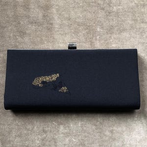 May include: A navy blue clutch with a silver clasp. The rectangular bag features a gold and black abstract design on the front. The clutch is likely made of a fabric material.