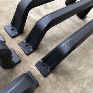 Peut inclure: Un ensemble de poignées de placard en métal noir mat présentant un design arqué et incurvé avec des plaques de montage plates à chaque extrémité. Chaque poignée comprend deux trous de vis pré-percés pour l'installation, avec une finition sombre texturée.