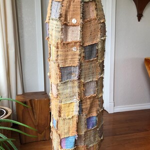 May include: A long patchwork skirt with a button closure. The skirt is made of different fabrics in shades of brown, beige, blue, and purple. The fabric has a frayed edge.