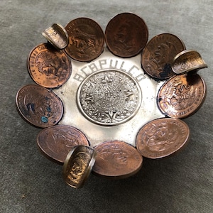 May include: A circular metal dish featuring a central silver-toned medallion with an intricate sun design and the text ACAPULCO. The rim is decorated with multiple copper-colored coins, some of which are bent upwards to form small curved edges.