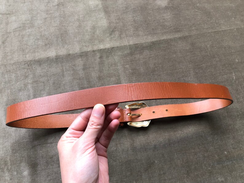 May include: A brown leather belt with a gold-toned buckle. The buckle has a decorative design.