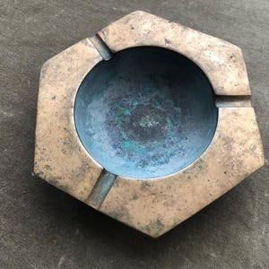 May include: A vintage hexagonal ashtray with a central circular depression. The ashtray is made of aged brass, showing signs of wear and a patina. The interior has a blue-green hue, and there are three grooves for resting cigarettes.