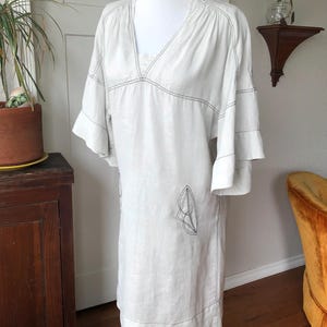 May include: A white linen dress with a v-neck and short sleeves. The dress has a simple design with black stitching details and a geometric pattern on the front.