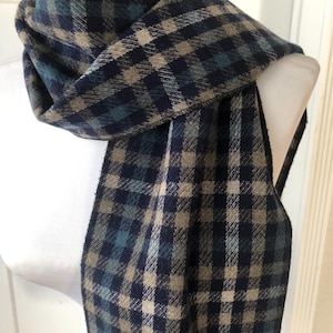 May include: A scarf featuring a plaid pattern in shades of navy blue, dark green, and tan. The fabric has a textured, woven appearance with a consistent grid design throughout the length of the accessory, draped around a white mannequin neck.
