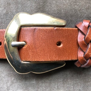 May include: A brown leather belt with a gold metal buckle and a braided leather end. The buckle is a simple, rounded design.
