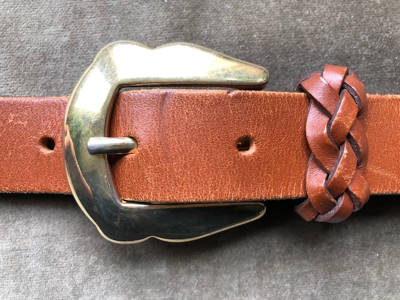 May include: A brown leather belt with a gold metal buckle and a braided leather end. The buckle is a simple, rounded design.