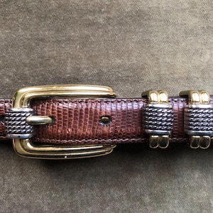 Puede incluir: Un cinturón de cuero marrón con una hebilla dorada y dos cierres de cinturón plateados y dorados. El cinturón tiene un patrón texturizado, similar a la piel de serpiente.