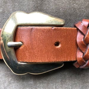 May include: A brown leather belt with a gold buckle and a braided leather end. The buckle is a simple, rounded design.