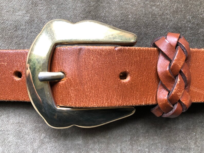 May include: A brown leather belt with a gold buckle and a braided leather end. The buckle is a simple, rounded design.