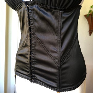 Pode incluir: Corpete de cetim preto com fecho frontal de gancho e ilh&oacute;s, com guarni&ccedil;&atilde;o decorativa e al&ccedil;as ajust&aacute;veis. O corpete tem cintas de liga.