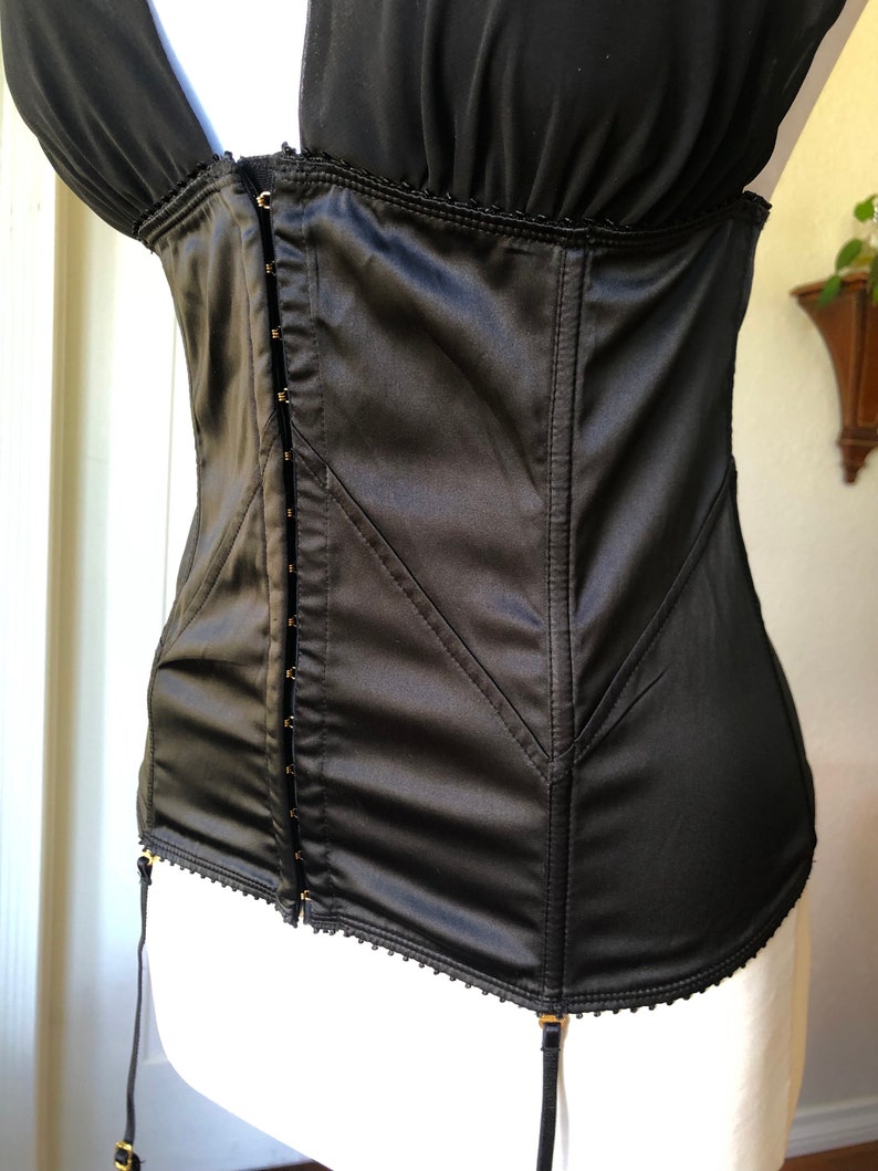 Pode incluir: Corpete de cetim preto com fecho frontal de gancho e ilh&oacute;s, com guarni&ccedil;&atilde;o decorativa e al&ccedil;as ajust&aacute;veis. O corpete tem cintas de liga.