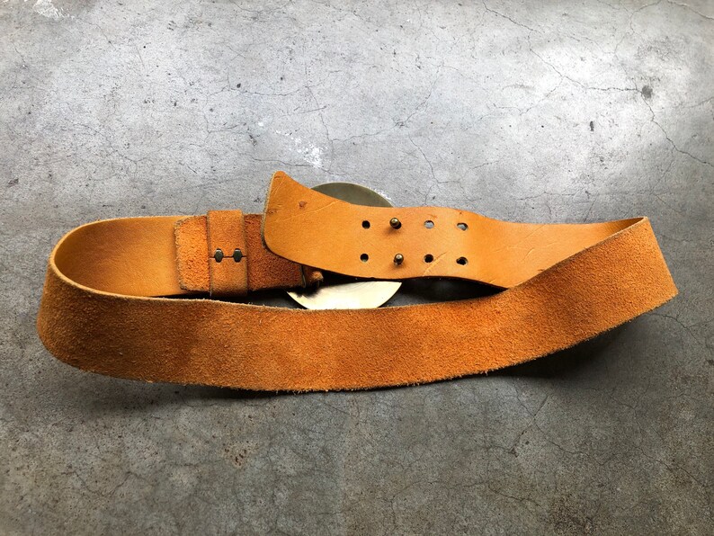 May include: A brown leather belt with a large, round, metal buckle. The buckle has a decorative design and is secured with small metal studs.