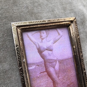 May include: A vintage photograph in a gold-colored metal frame. The photo features a woman in a swimsuit posing on a beach. The frame has a decorative, etched border. The image has a sepia tone.