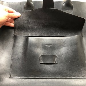 Puede incluir: Bolso tote de cuero negro con un bolsillo grande con solapa y un bolsillo m&aacute;s peque&ntilde;o en el interior. El bolso tiene una etiqueta de cuero que dice "Made in USA".