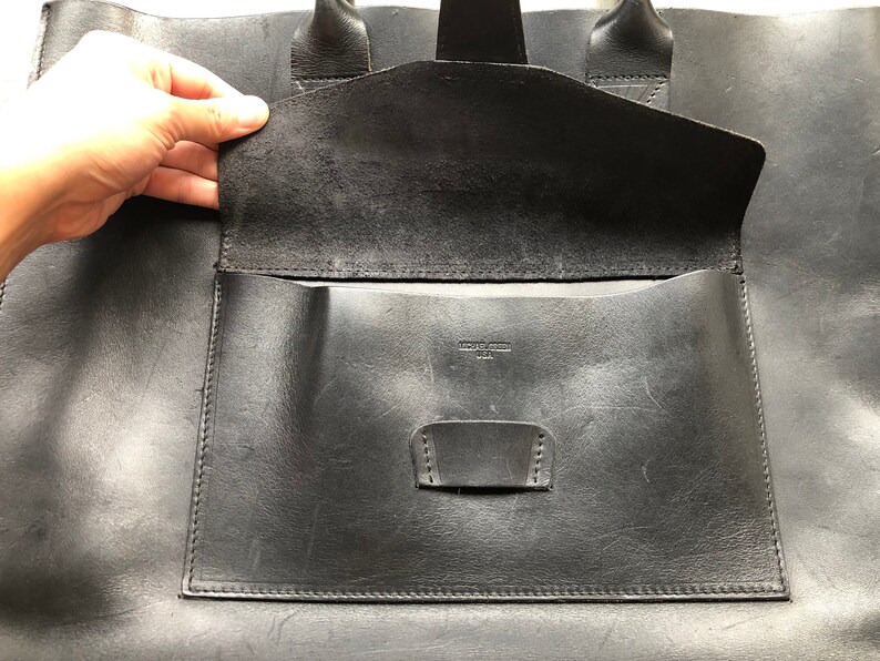 Puede incluir: Bolso tote de cuero negro con un bolsillo grande con solapa y un bolsillo m&aacute;s peque&ntilde;o en el interior. El bolso tiene una etiqueta de cuero que dice "Made in USA".