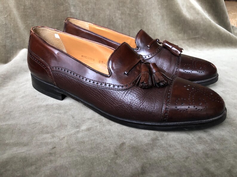 May include: A pair of brown leather tassel loafers with a wingtip design. The shoes have a brogue pattern and a tassel detail on the vamp.