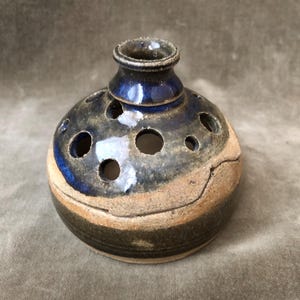 May include: A handcrafted ceramic vase with a round body and a small, cylindrical neck. The vase features a dark blue glaze on the neck and upper body, with a speckled, tan-colored base. It has several circular openings.